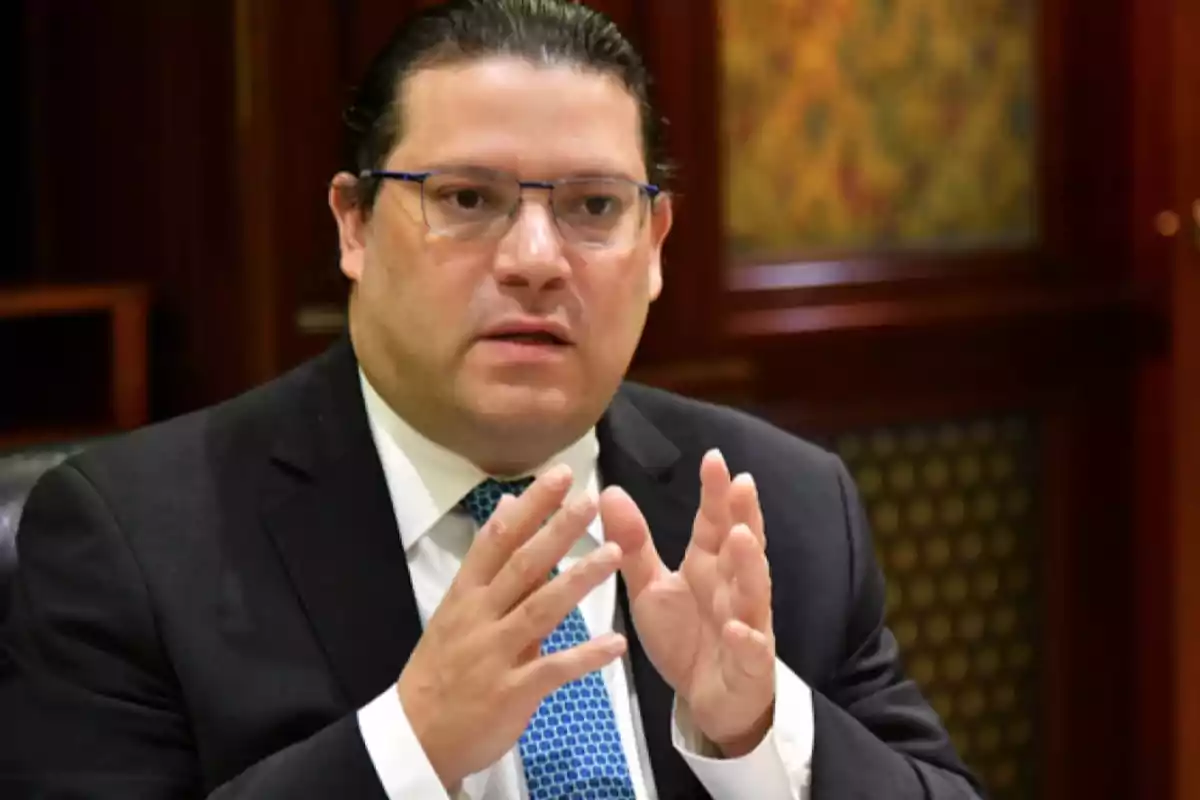 Hombre con gafas y traje oscuro hablando y gesticulando con las manos en un entorno formal