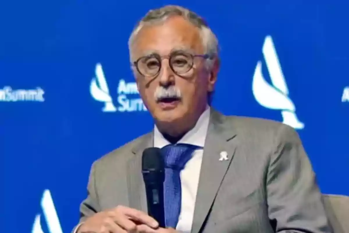Gray-haired man with a mustache, wearing glasses and a gray suit, speaking at an event with a blue background Gray-haired man with a mustache, wearing glasses and a gray suit, speaking at an event with a blue background