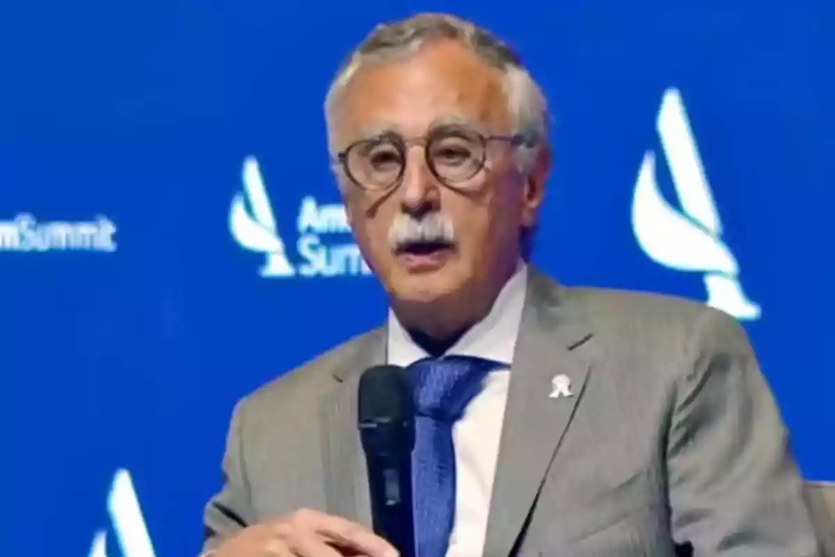 Hombre mayor con gafas y bigote usando traje gris y corbata azul mientras sostiene un micrófono frente a un fondo azul con logotipos blancos Hombre mayor con gafas y bigote usando traje gris y corbata azul mientras sostiene un micrófono frente a un fondo azul con logotipos blancos