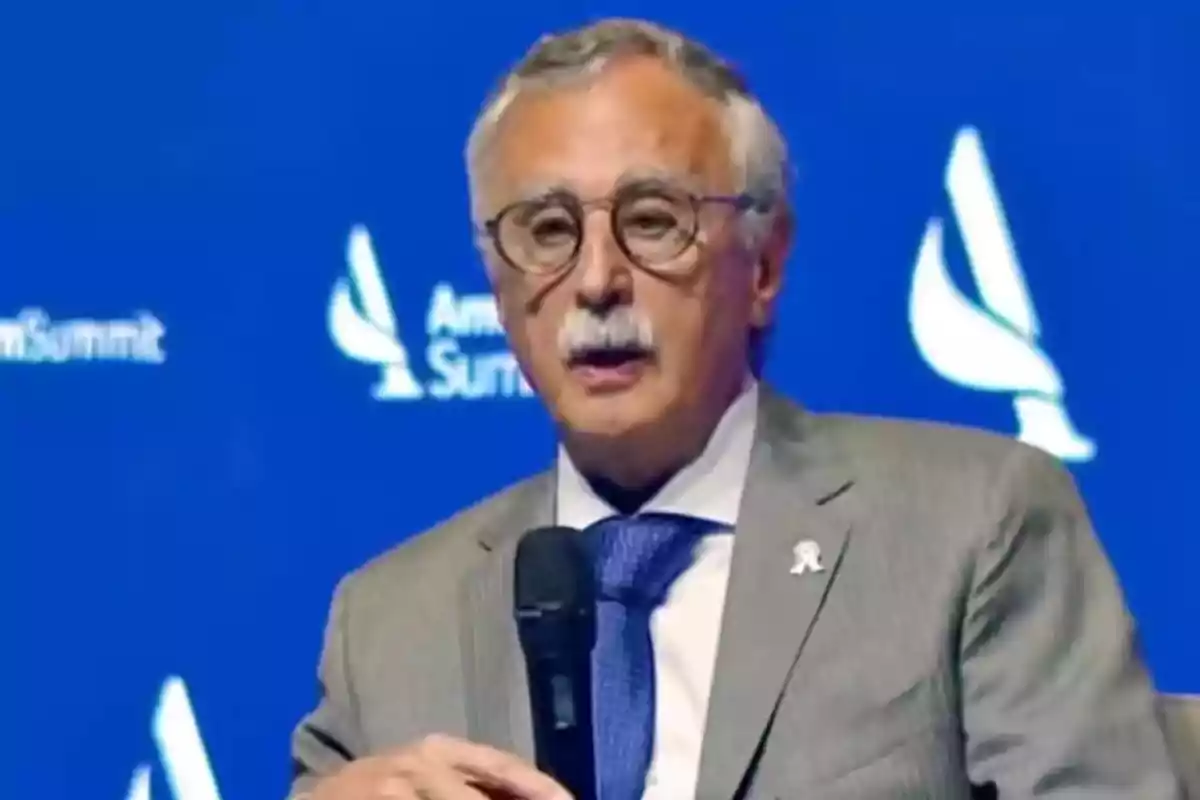 Hombre mayor con gafas y bigote usando traje gris y corbata azul mientras sostiene un micrófono frente a un fondo azul con logotipos blancos Hombre mayor con gafas y bigote usando traje gris y corbata azul mientras sostiene un micrófono frente a un fondo azul con logotipos blancos
