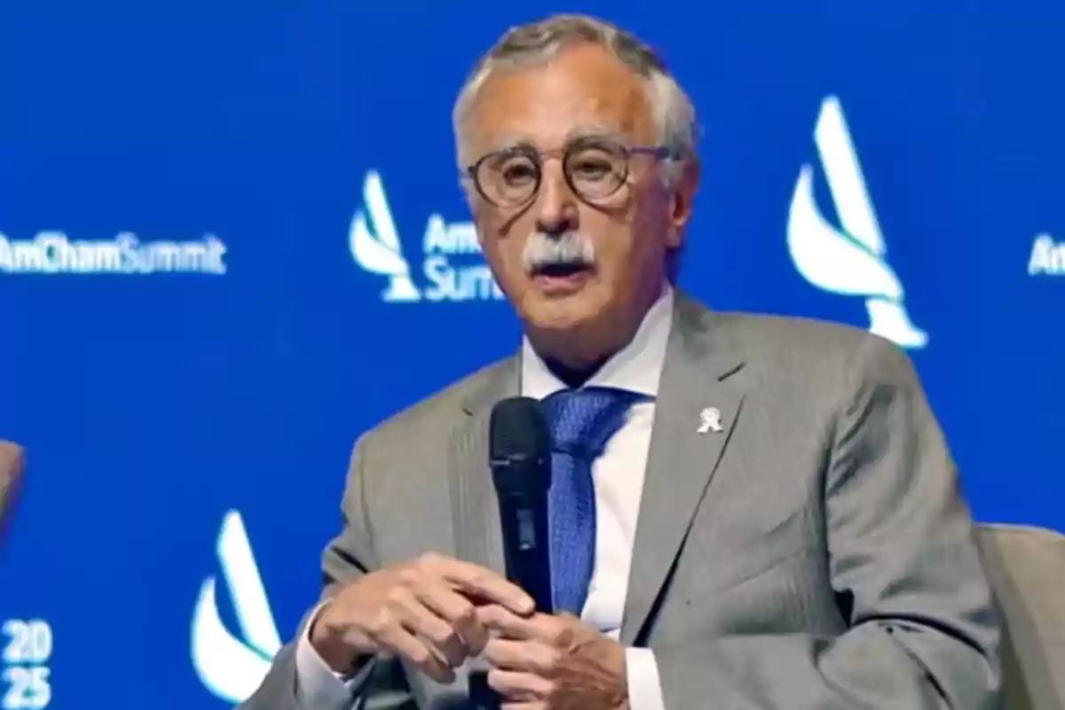 Hombre de cabello canoso y bigote con gafas y traje gris hablando en un evento con fondo azul que tiene el logo de AmCham Summit