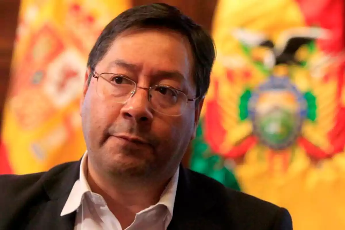Man with glasses and a serious expression in front of colorful, blurred flags in the background
