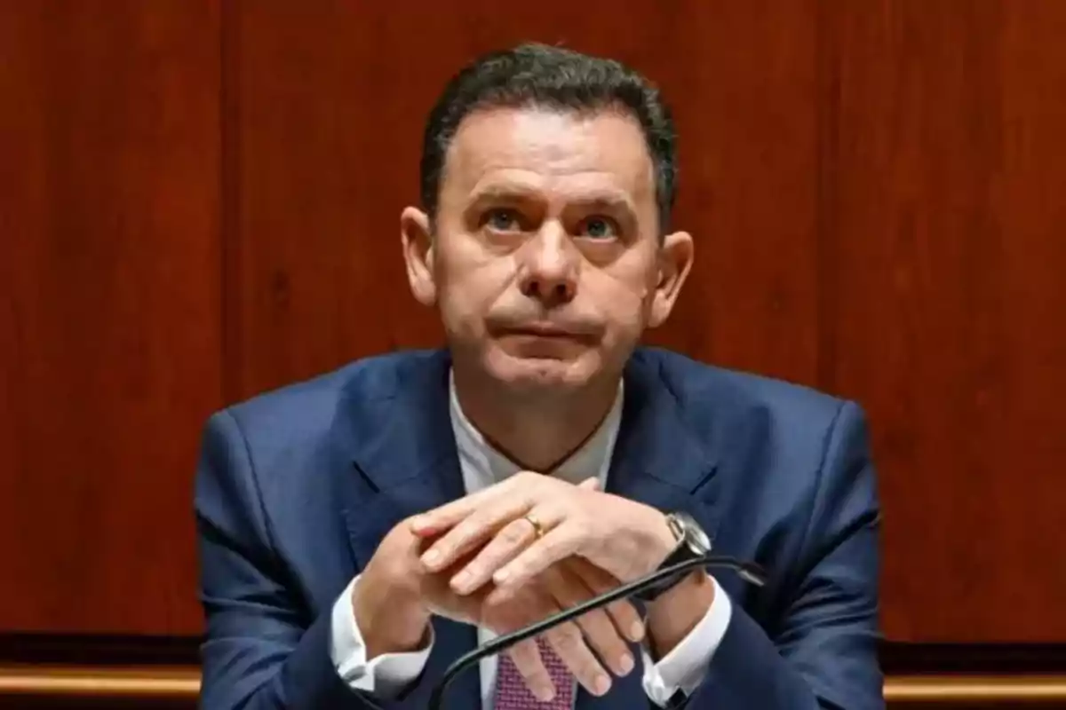 Man in a blue suit sitting with his hands clasped and a serious expression in front of a wooden background Man in a blue suit sitting with his hands clasped and a serious expression in front of a wooden background