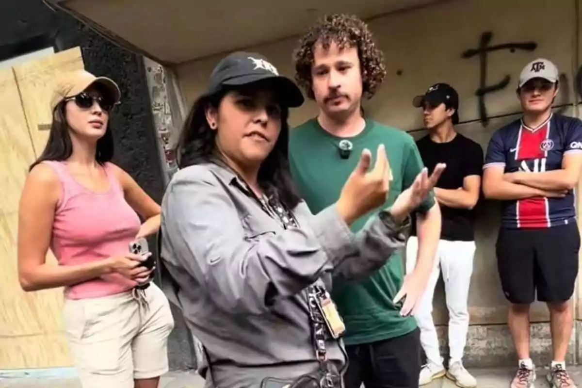 Group of five people standing on the street, a woman in front gesturing while the others watch, some with their arms crossed and wearing caps and casual clothes Group of five people standing on the street, a woman in front gesturing while the others watch, some with their arms crossed and wearing caps and casual clothes