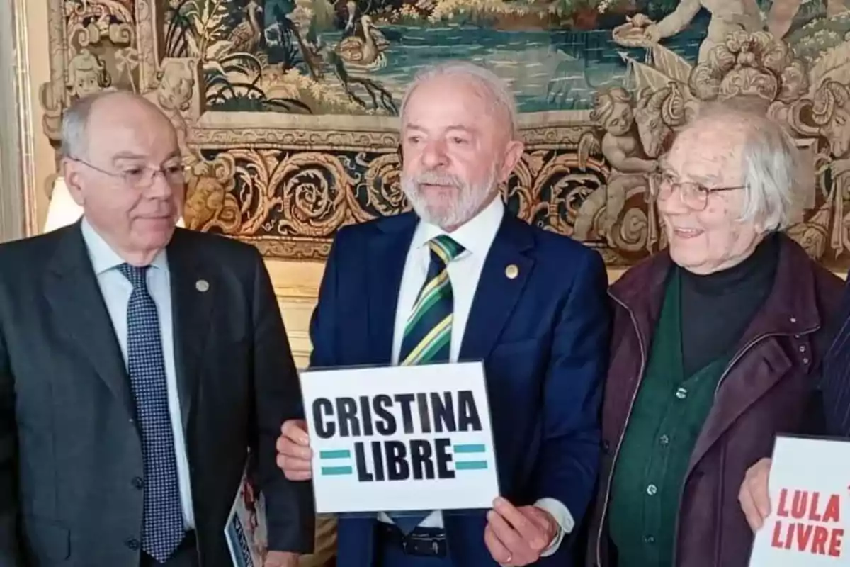 Tres personas mayores posan sonrientes en un ambiente elegante, una de ellas sostiene un cartel que dice CRISTINA LIBRE y otra sostiene un cartel que dice LULA LIVRE