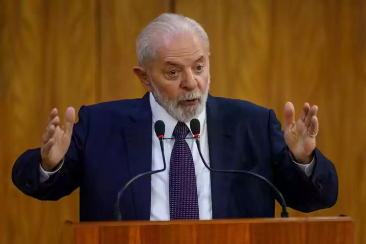Hombre de cabello canoso y barba hablando en un podio con micrófonos y gesticulando con las manos, fondo de madera