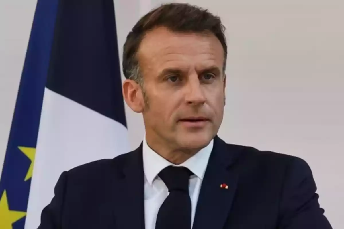 Man in a dark suit and black tie speaking in front of a flag with blue, white, and yellow colors.