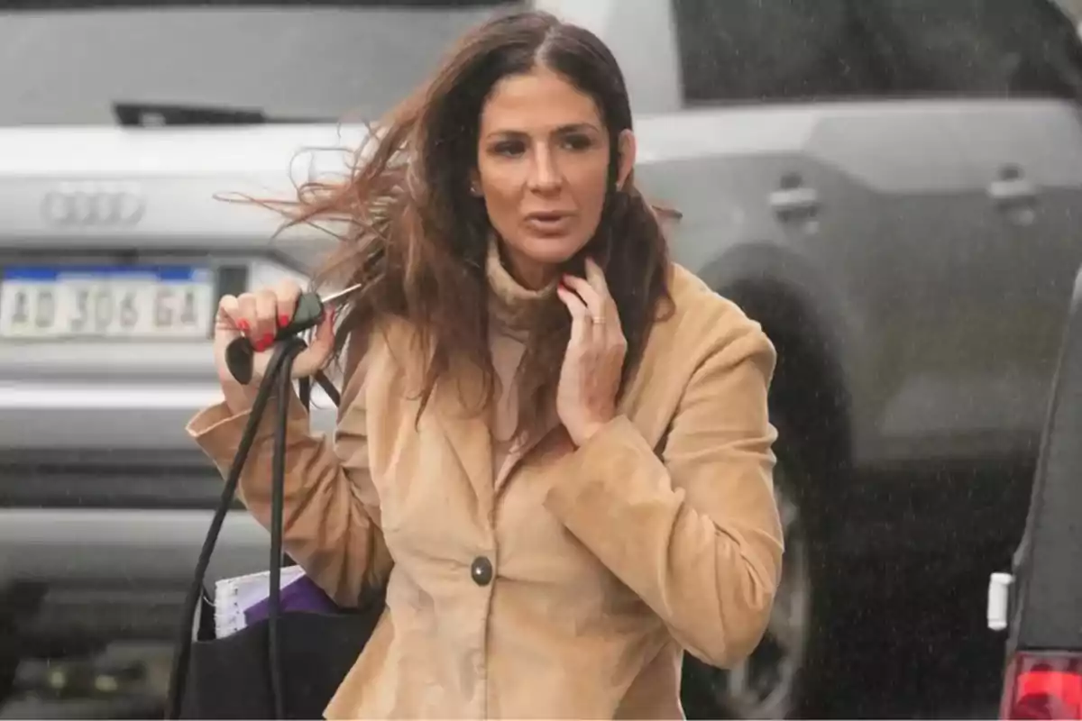 Mujer de cabello largo y castaño con saco beige caminando en la calle mientras sostiene una bolsa negra y unas llaves en un día lluvioso