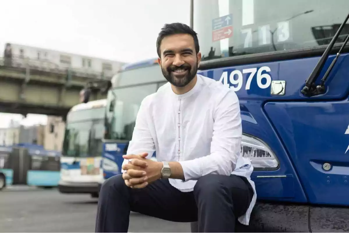 Hombre sonriente con barba y camisa blanca sentado frente a un autobús azul en un entorno urbano Hombre sonriente con barba y camisa blanca sentado frente a un autobús azul en un entorno urbano