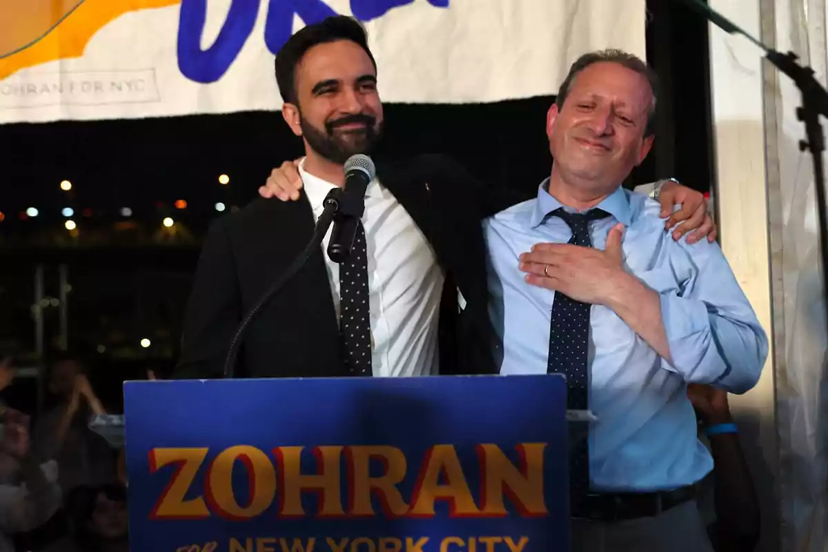 Two men smile as they embrace on a stage during a political campaign event, one of them standing behind a podium with a sign that reads "ZOHARAN FOR NEW YORK CITY."