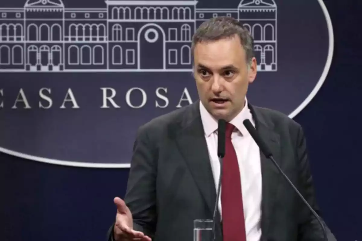 Hombre de traje y corbata roja hablando en una conferencia de prensa frente a un fondo con el dibujo de un edificio y las palabras Casa Rosada Hombre de traje y corbata roja hablando en una conferencia de prensa frente a un fondo con el dibujo de un edificio y las palabras Casa Rosada