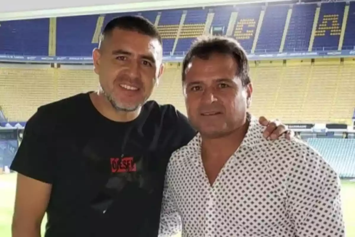Dos hombres posan juntos sonriendo en el interior de un estadio de fútbol con gradas amarillas y azules de fondo Dos hombres posan juntos sonriendo en el interior de un estadio de fútbol con gradas amarillas y azules de fondo