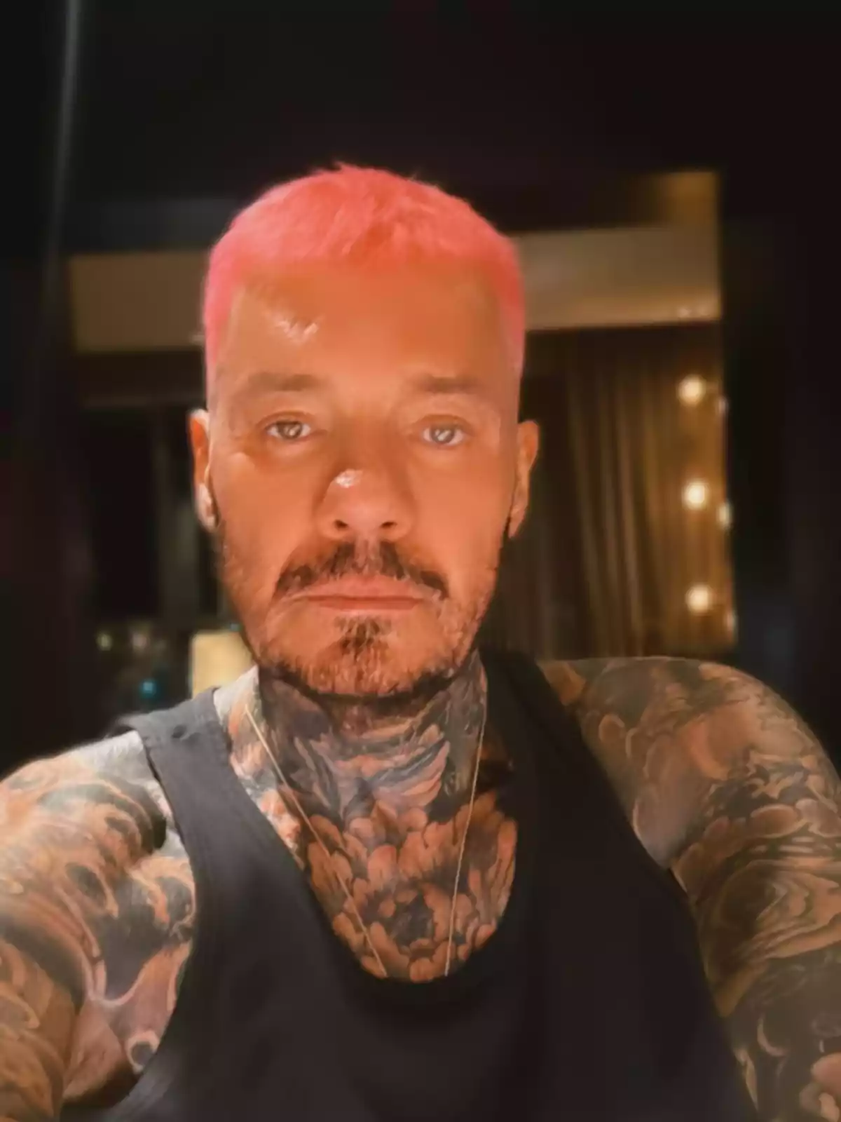Hombre con cabello corto teñido de rosa, barba, tatuajes en el cuello y brazos, usando una camiseta sin mangas negra en un ambiente interior con luz tenue Hombre con cabello corto teñido de rosa, barba, tatuajes en el cuello y brazos, usando una camiseta sin mangas negra en un ambiente interior con luz tenue
