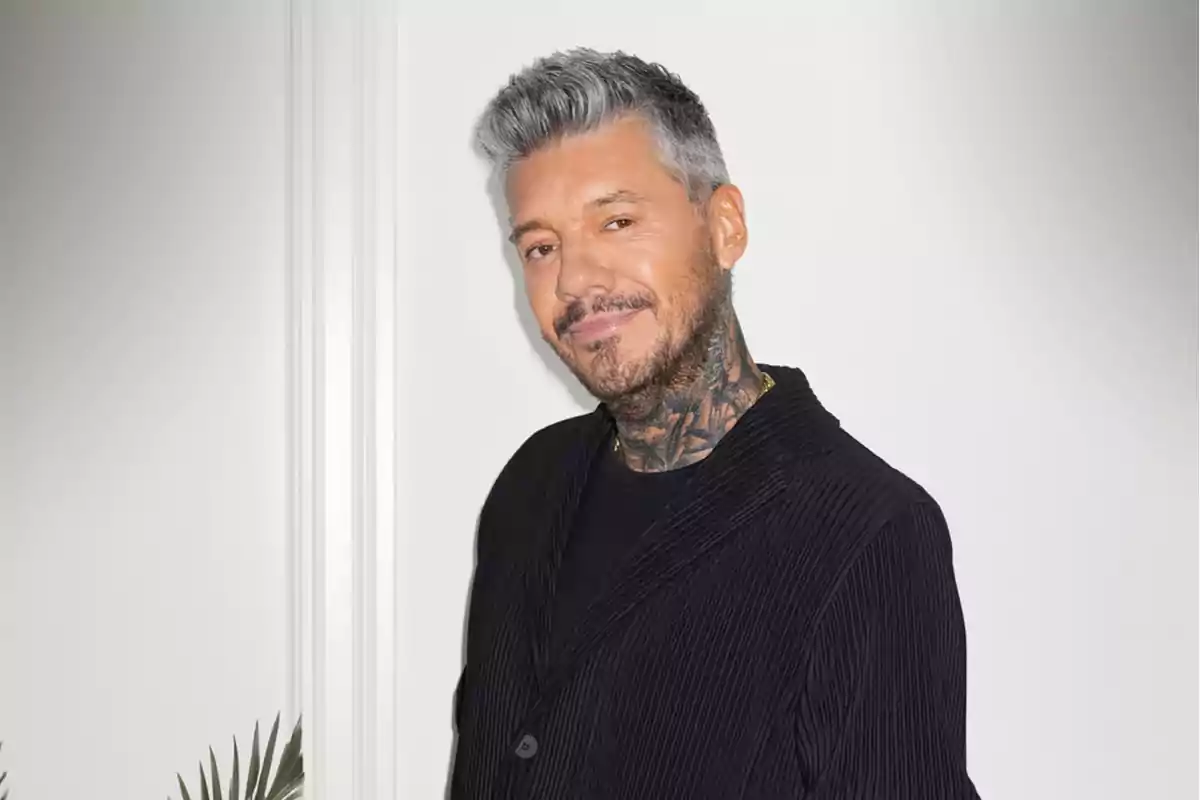 Hombre de cabello canoso y barba con tatuajes en el cuello, vistiendo un saco negro y sonriendo frente a una pared blanca