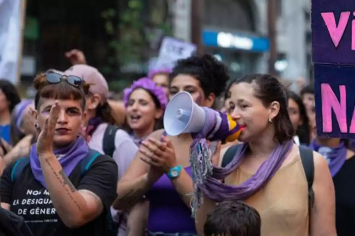 Women marching in a feminist demonstration, one of them using a megaphone and several wearing purple scarves and accessories.