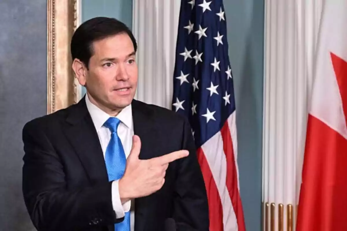 Man in a dark suit and blue tie pointing to the side with United States and Canadian flags in the background