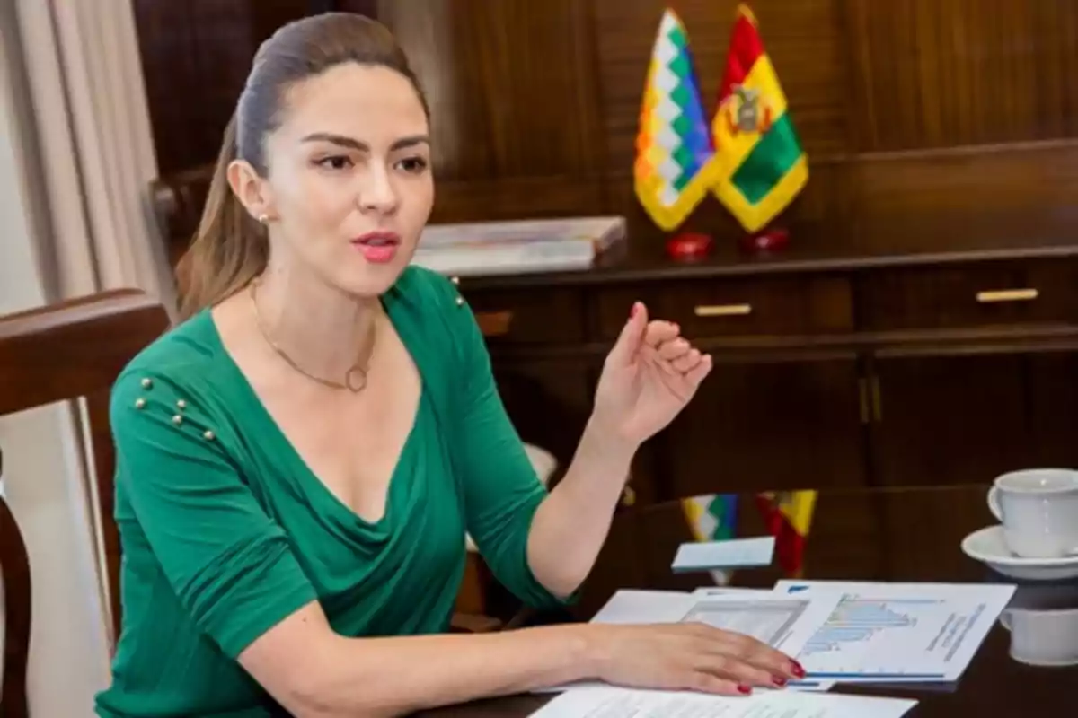 Mujer sentada en una oficina con documentos y gráficos sobre la mesa, hablando y gesticulando, al fondo se ven dos banderas de Bolivia Mujer sentada en una oficina con documentos y gráficos sobre la mesa, hablando y gesticulando, al fondo se ven dos banderas de Bolivia