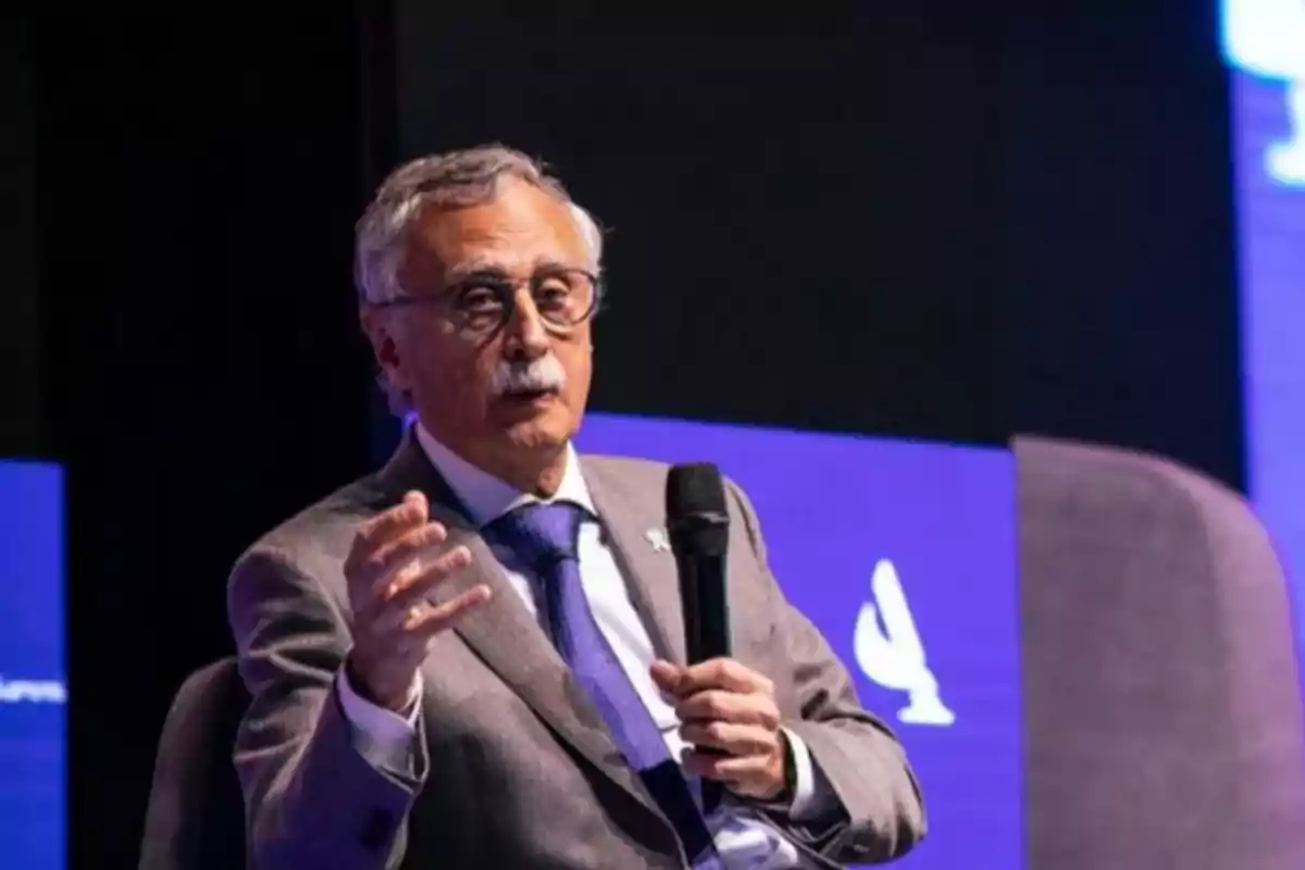 Hombre mayor con gafas y bigote hablando mientras sostiene un micrófono en un escenario con fondo azul