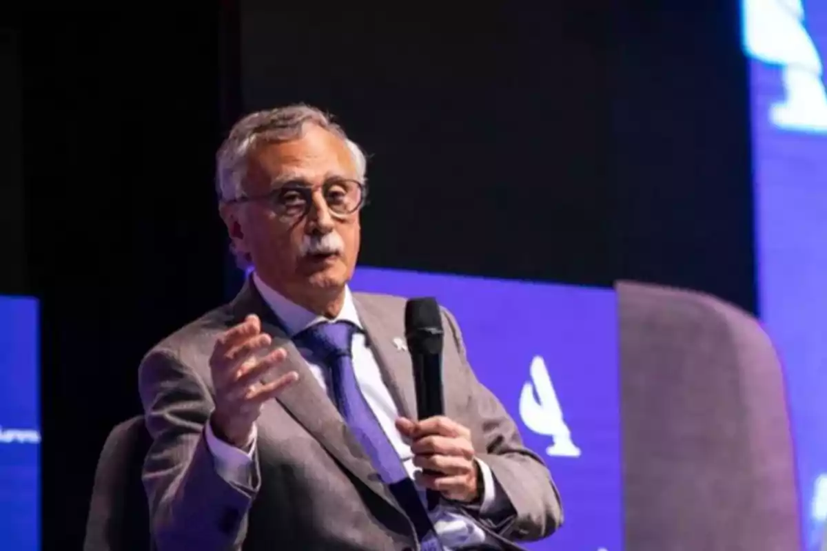 Hombre mayor con gafas y bigote hablando mientras sostiene un micrófono en un escenario con fondo azul