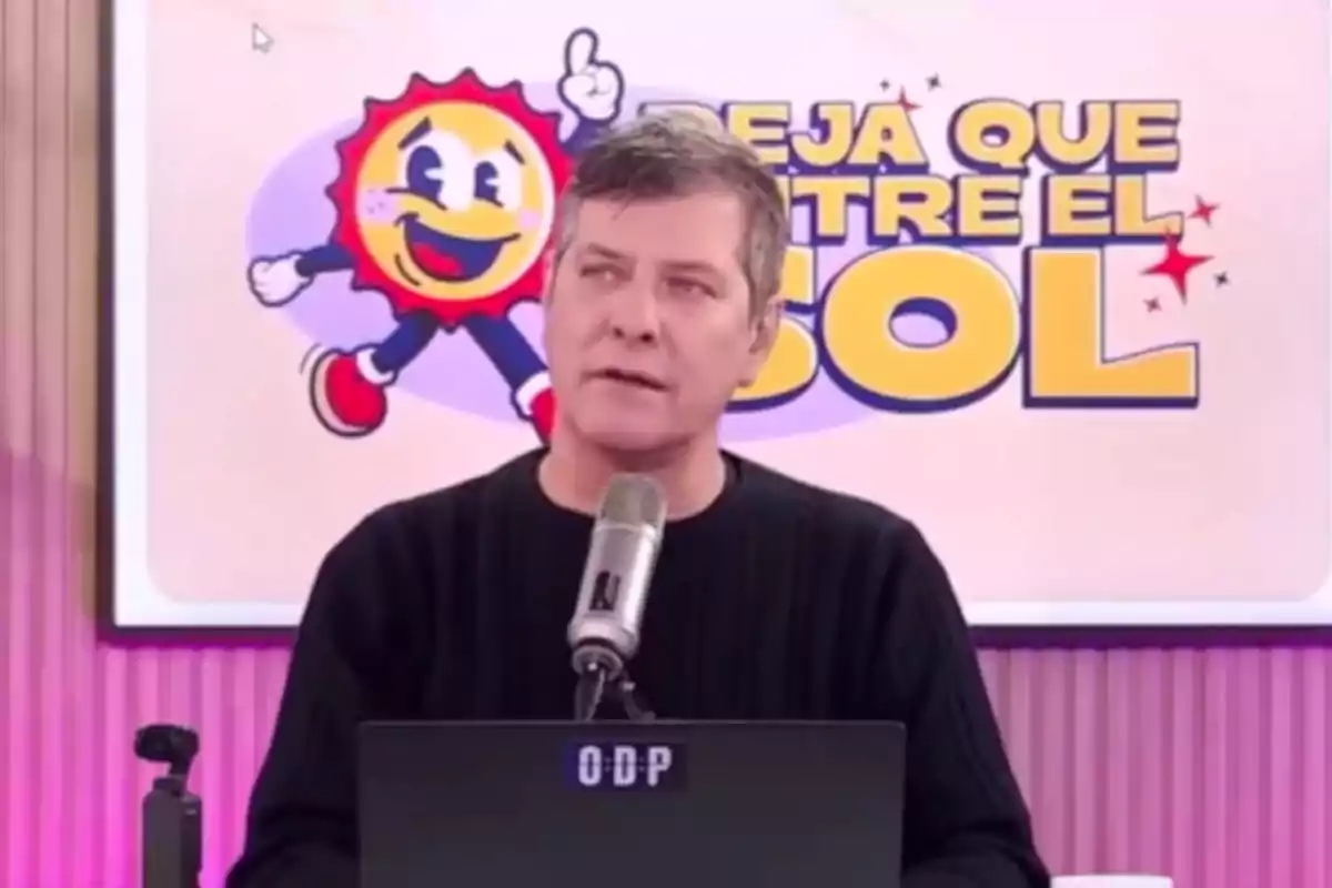 Hombre de cabello corto y suéter oscuro hablando frente a un micrófono con una laptop, al fondo un cartel colorido con un sol sonriente y letras grandes amarillas. Hombre de cabello corto y suéter oscuro hablando frente a un micrófono con una laptop, al fondo un cartel colorido con un sol sonriente y letras grandes amarillas.