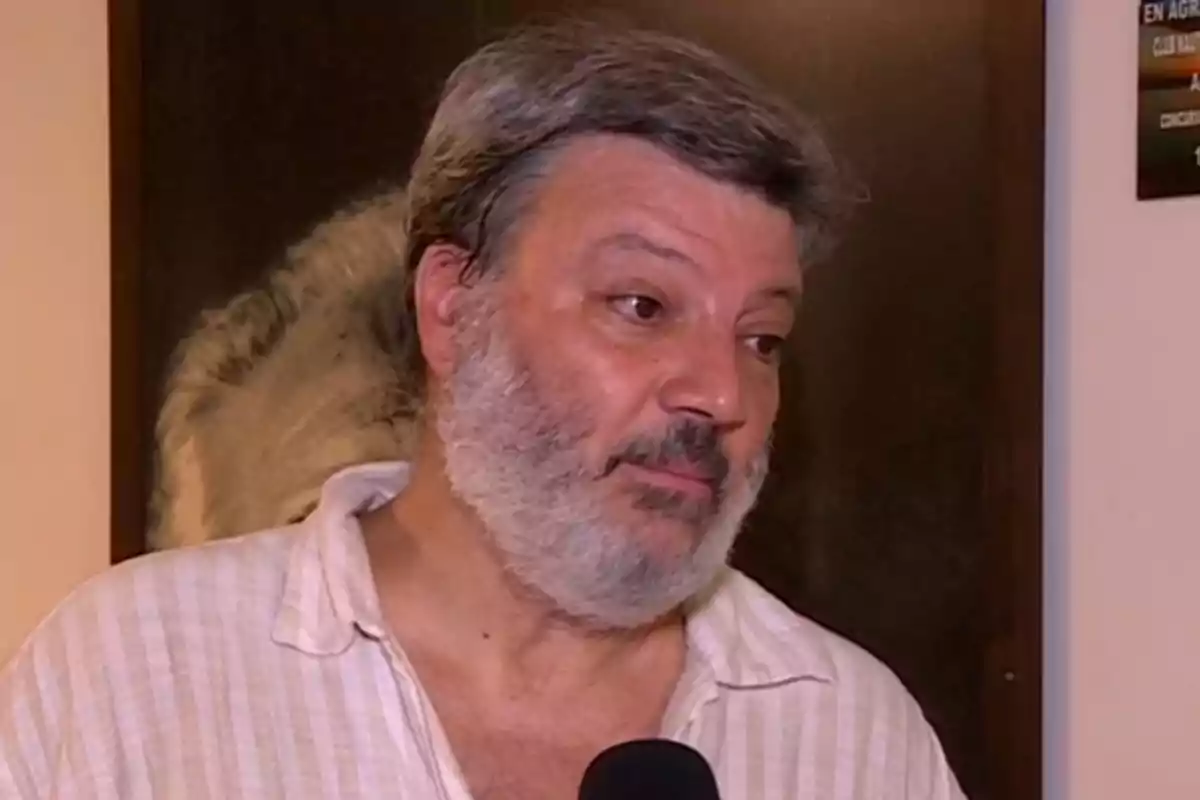 Hombre de cabello y barba canosos con camisa clara siendo entrevistado con un micrófono en un interior Hombre de cabello y barba canosos con camisa clara siendo entrevistado con un micrófono en un interior