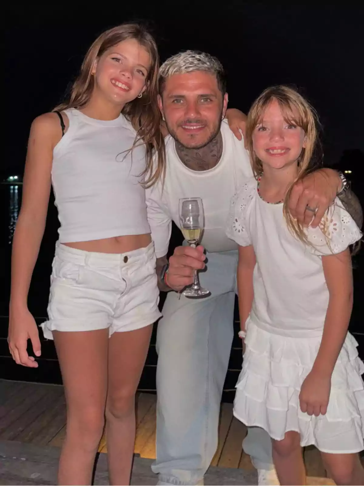 A smiling man with two girls by his side, all dressed in white and posing together outdoors at night. A smiling man with two girls by his side, all dressed in white and posing together outdoors at night.