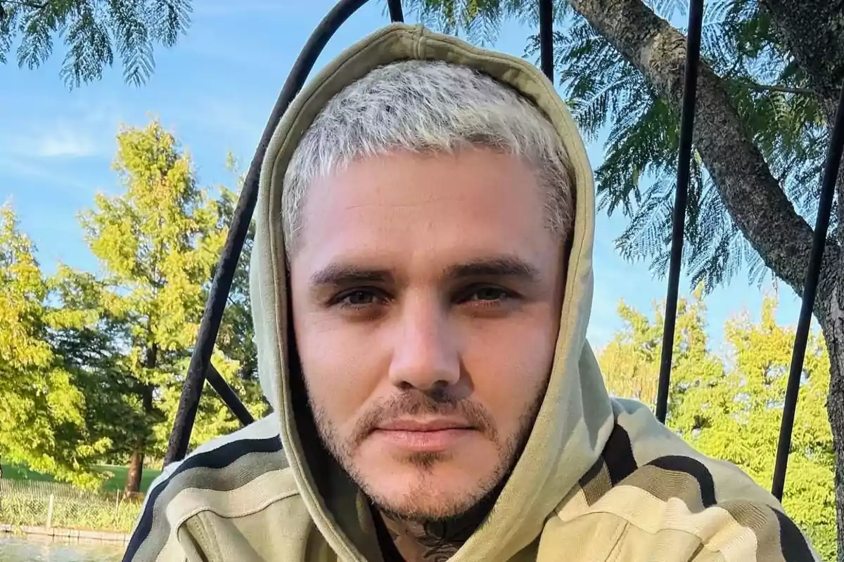 Joven con cabello corto y rubio usando sudadera con capucha beige, sentado al aire libre con árboles y cielo azul de fondo