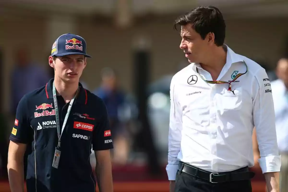 Two men are talking in a Formula 1 paddock; one is wearing a Red Bull uniform and the other is dressed in a white Mercedes jersey. Two men are talking in a Formula 1 paddock; one is wearing a Red Bull uniform and the other is dressed in a white Mercedes jersey.
