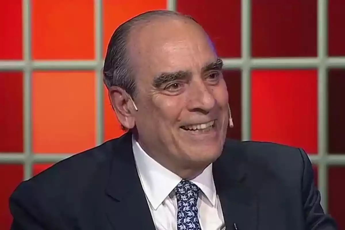 Man in a dark suit and tie smiles while participating in a television show with a background of red and orange squares. Man in a dark suit and tie smiles while participating in a television show with a background of red and orange squares.