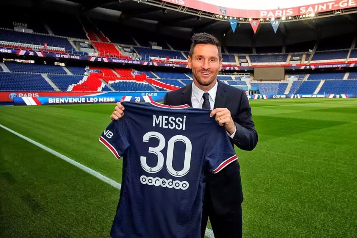 Un futbolista sonriente sostiene una camiseta azul con el número 30 y su nombre en un estadio vacío Un futbolista sonriente sostiene una camiseta azul con el número 30 y su nombre en un estadio vacío