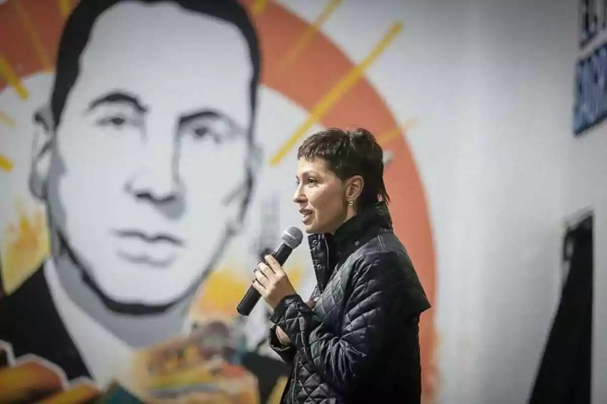 Mujer hablando por micrófono frente a un mural de un rostro masculino estilizado Mujer hablando por micrófono frente a un mural de un rostro masculino estilizado