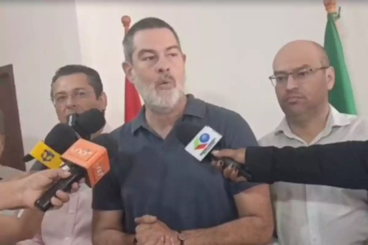 A bearded man in a blue t-jersey speaks in front of several microphones while two people stand behind him in the background.