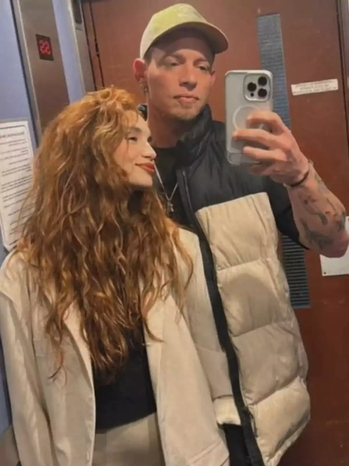 Una pareja se toma una selfie en un ascensor, ambos lucen relajados y sonrientes, el hombre sostiene el teléfono mientras la mujer lo mira. Una pareja se toma una selfie en un ascensor, ambos lucen relajados y sonrientes, el hombre sostiene el teléfono mientras la mujer lo mira.