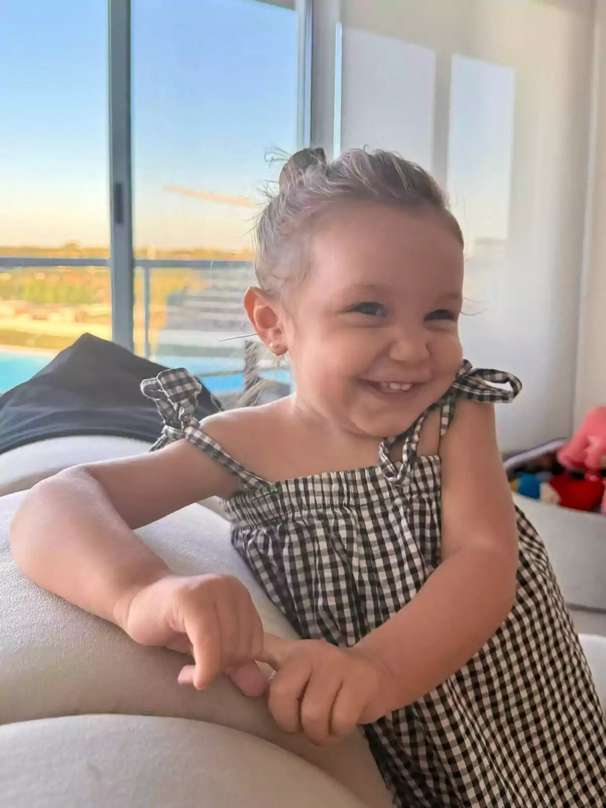 Niña pequeña sonriendo con un vestido de cuadros en una sala iluminada por la luz natural que entra por una ventana grande Niña pequeña sonriendo con un vestido de cuadros en una sala iluminada por la luz natural que entra por una ventana grande