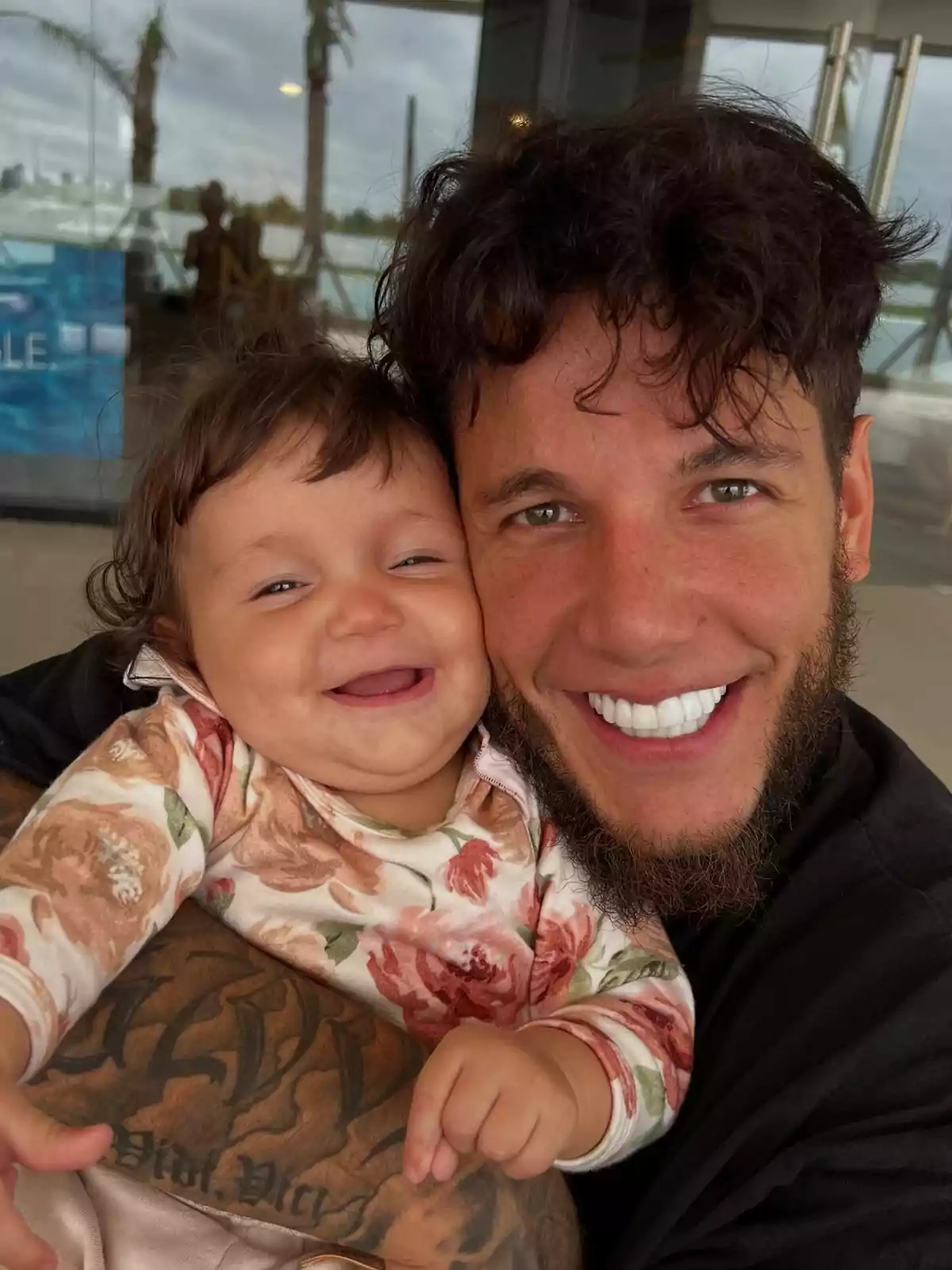Hombre con barba y cabello rizado sonríe abrazando a una bebé que también sonríe, ambos lucen felices y están en un ambiente exterior con palmeras al fondo Hombre con barba y cabello rizado sonríe abrazando a una bebé que también sonríe, ambos lucen felices y están en un ambiente exterior con palmeras al fondo
