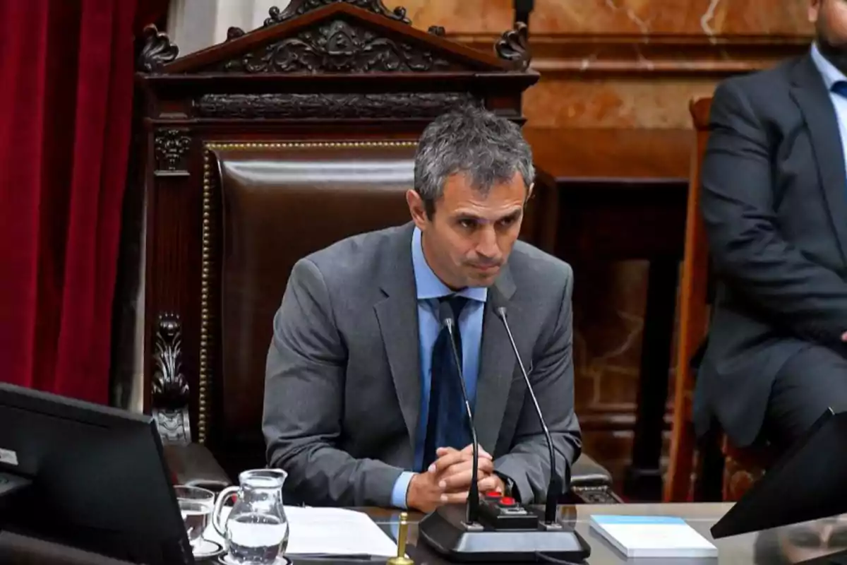 Hombre de traje gris sentado en una silla grande de madera en un recinto oficial con micrófonos frente a él y documentos sobre la mesa Hombre de traje gris sentado en una silla grande de madera en un recinto oficial con micrófonos frente a él y documentos sobre la mesa
