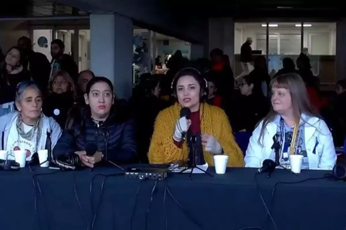 Cuatro mujeres sentadas en una mesa con micrófonos y vasos desechables, participando en una conferencia o panel frente a un grupo de personas en un ambiente interior.