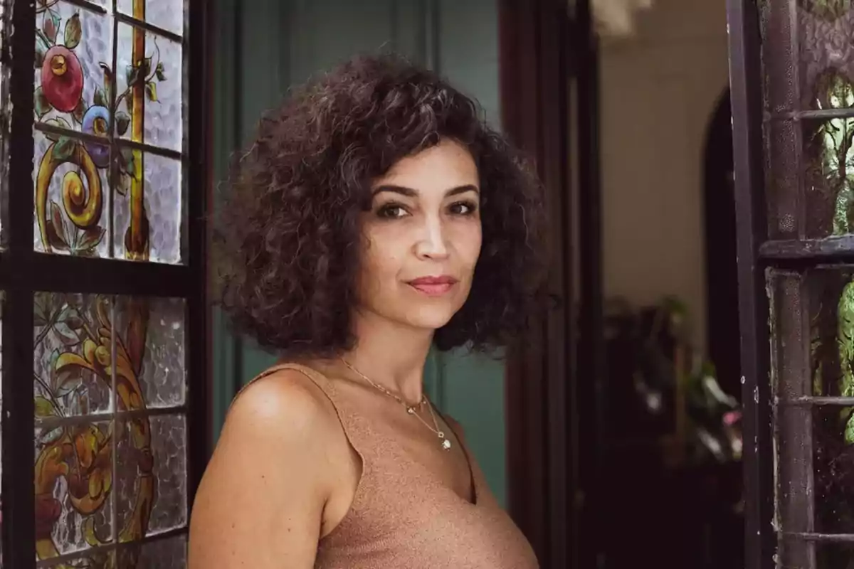 Mujer de cabello rizado y oscuro con vestido sin mangas color marrón claro posando junto a una puerta de vidrio decorado Mujer de cabello rizado y oscuro con vestido sin mangas color marrón claro posando junto a una puerta de vidrio decorado