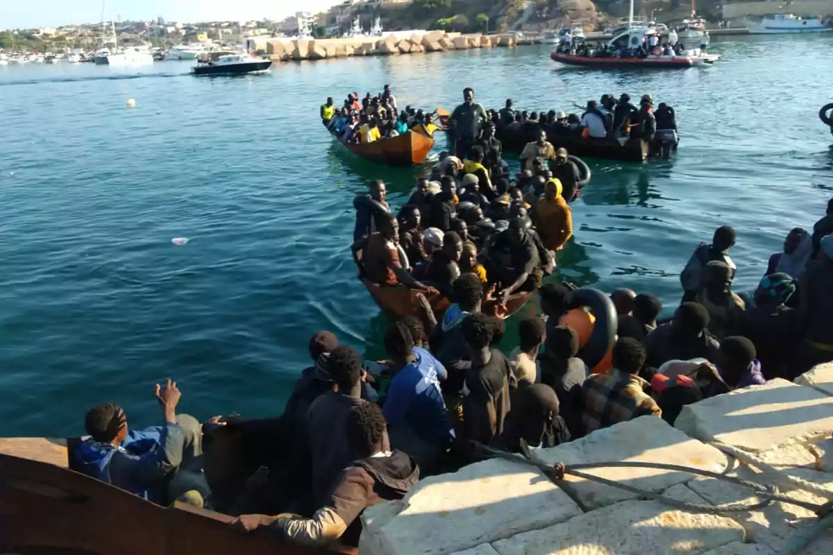 Varias embarcaciones llenas de personas migrantes llegan a un puerto mientras otras embarcaciones y barcos se ven al fondo Varias embarcaciones llenas de personas migrantes llegan a un puerto mientras otras embarcaciones y barcos se ven al fondo