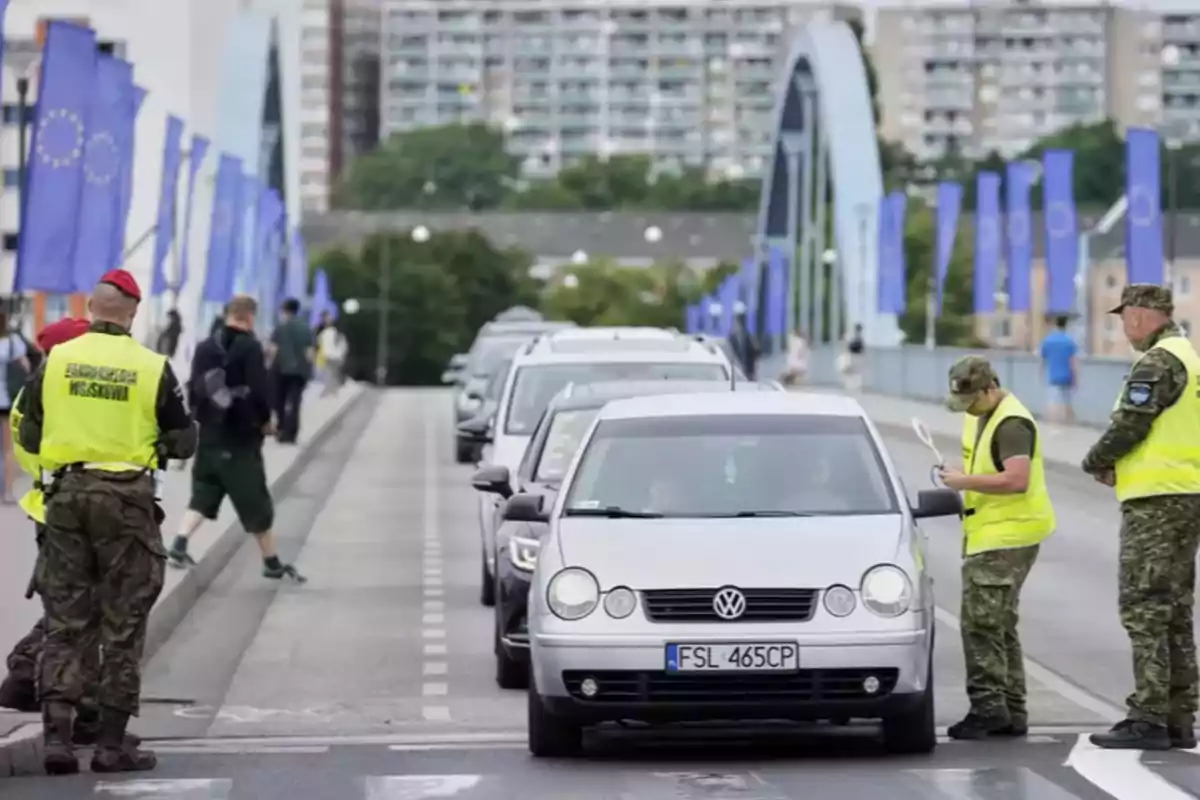 Control de vehículos en un puente con soldados revisando documentos y banderas de la Unión Europea a los lados Control de vehículos en un puente con soldados revisando documentos y banderas de la Unión Europea a los lados
