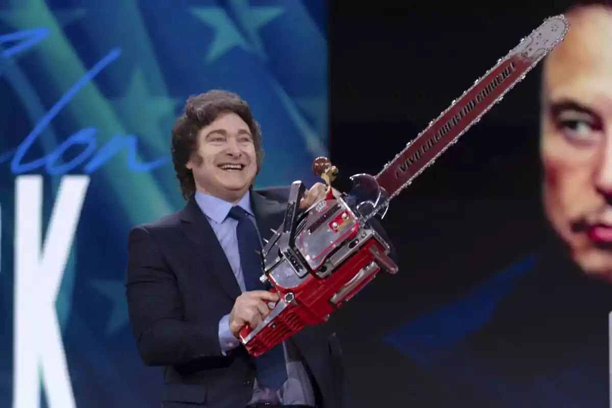 Hombre sonriente con traje sosteniendo una motosierra roja en un escenario con fondo azul y una cara parcialmente visible a la derecha