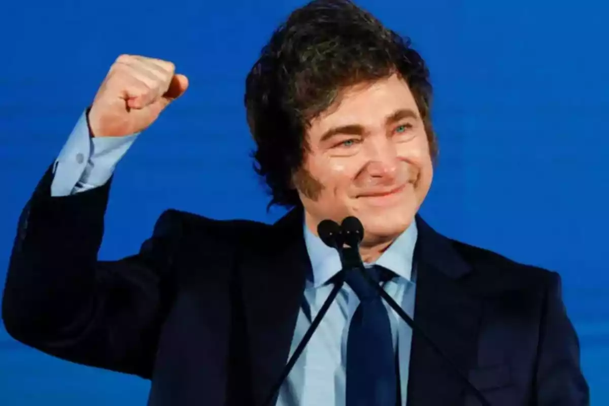 Dark-haired man in a suit and blue tie raising his fist and smiling in front of a microphone and a blue background