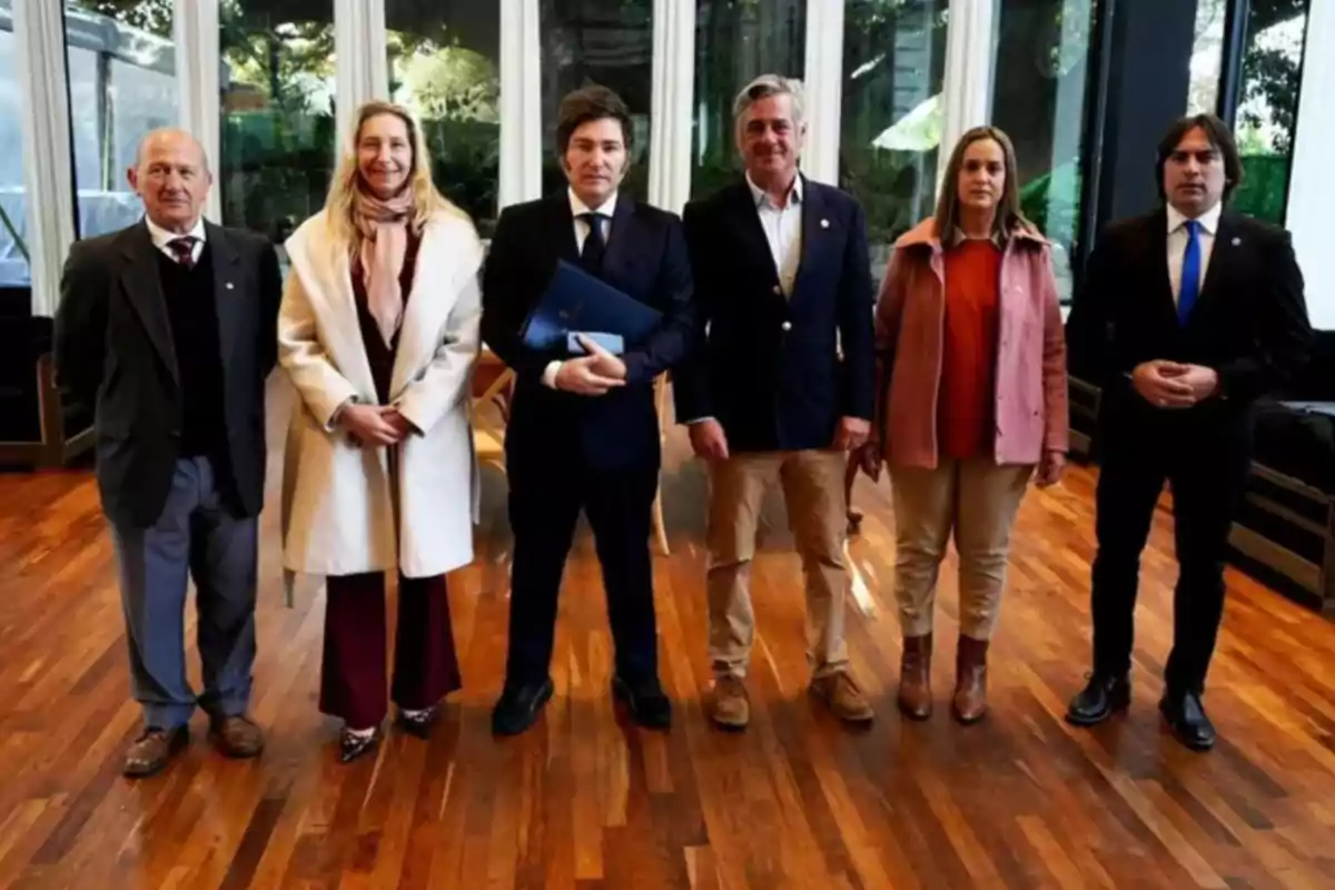 Seis personas posan de pie en fila dentro de un salón con piso de madera y grandes ventanales al fondo, todas vestidas de manera formal.