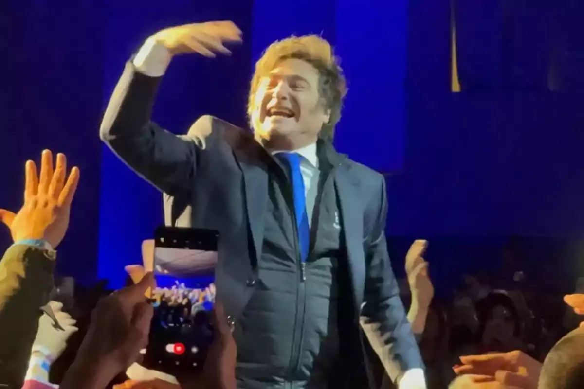 Smiling man in a blue suit and tie greeting a crowd around him while several people raise their hands and one holds a mobile phone recording the moment Smiling man in a blue suit and tie greeting a crowd around him while several people raise their hands and one holds a mobile phone recording the moment
