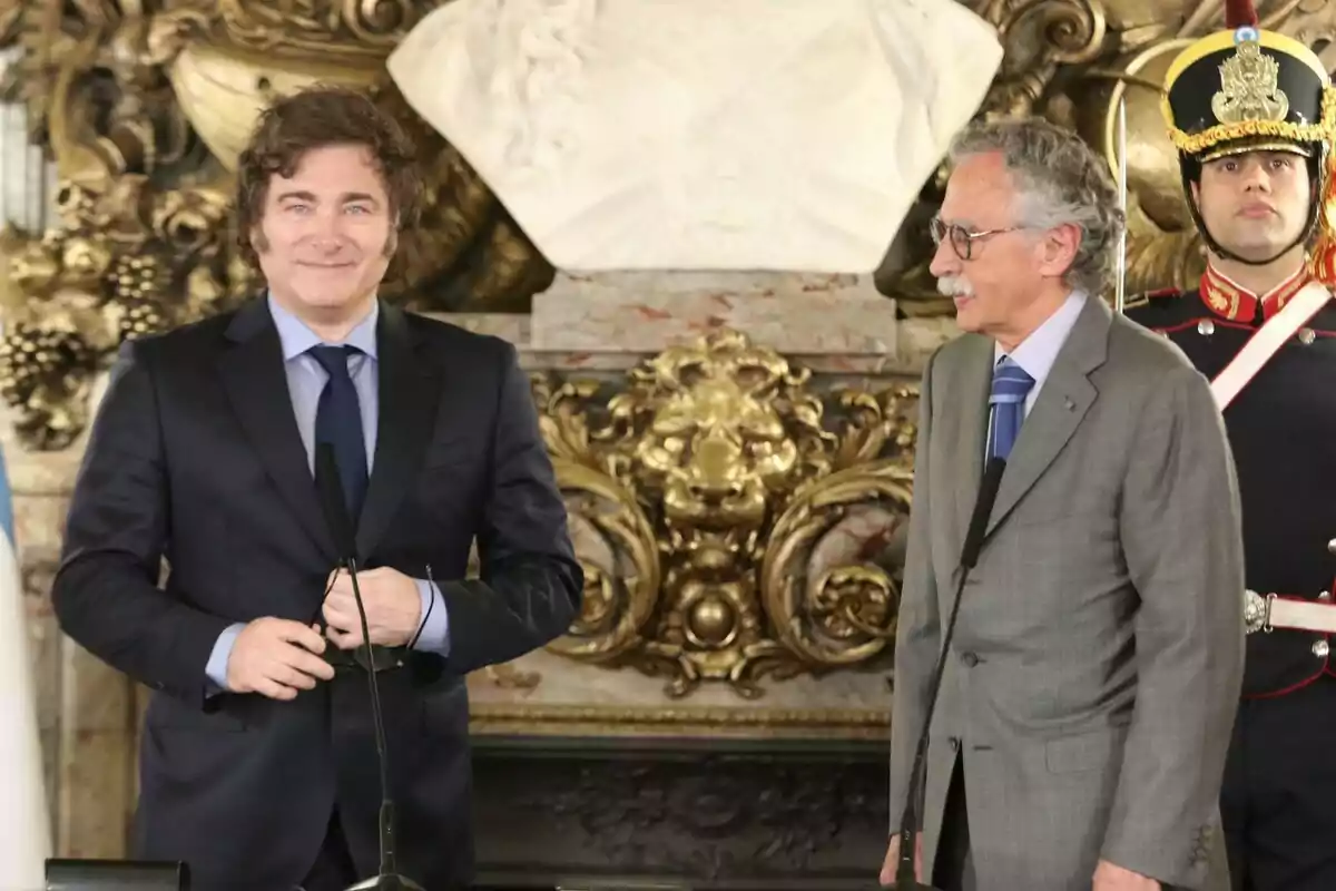 Two men in suits are talking in front of a decorative golden backdrop and a white statue, while a guard in ceremonial uniform stands behind them.