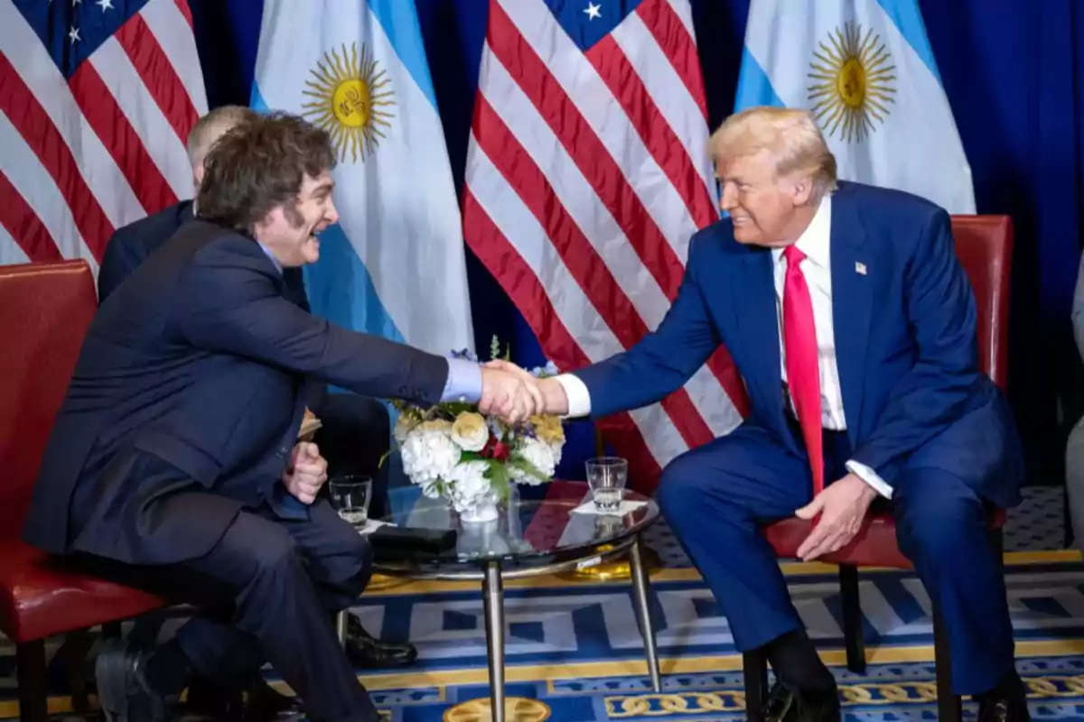 Dos hombres con traje se dan la mano sentados frente a una mesa pequeña con banderas de Argentina y Estados Unidos de fondo Dos hombres con traje se dan la mano sentados frente a una mesa pequeña con banderas de Argentina y Estados Unidos de fondo