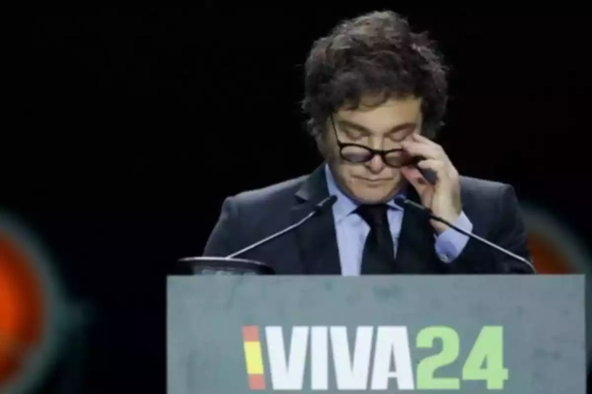 Curly-haired man with glasses adjusting his frames while speaking at a podium with the text VIVA24 Curly-haired man with glasses adjusting his frames while speaking at a podium with the text VIVA24