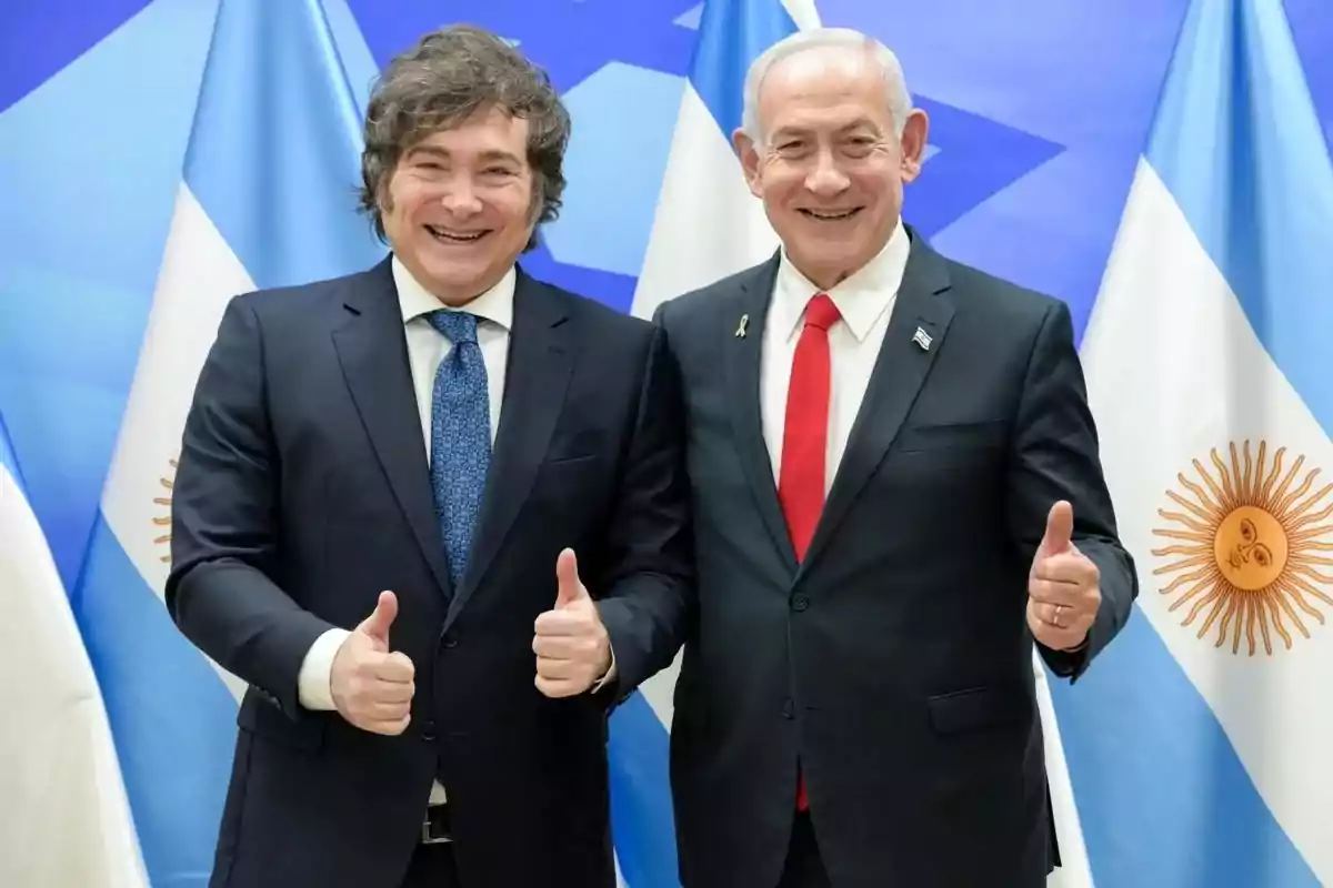 Two men in suits smile and pose together giving a thumbs-up gesture in front of the flags of Argentina and another blue and white flag.