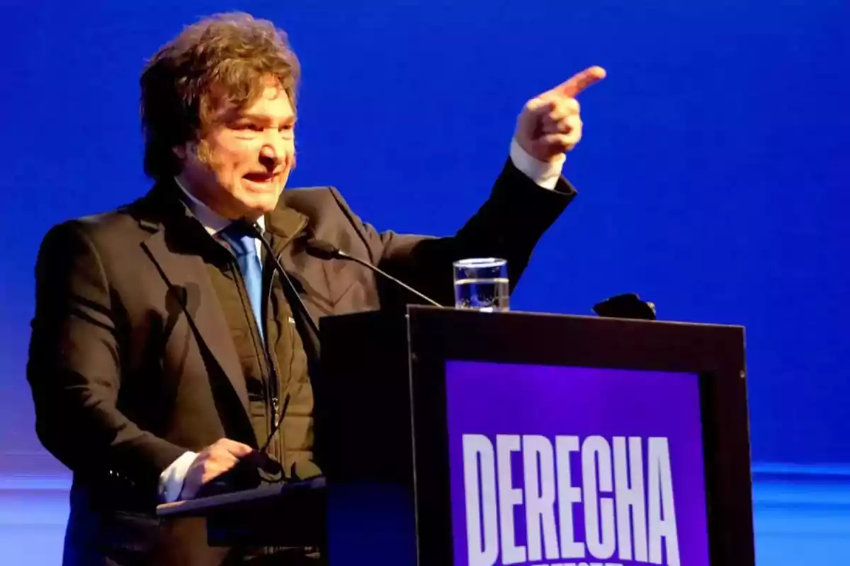 Hombre de cabello rizado y traje oscuro hablando en un podio con fondo azul y un cartel que dice DERECHA Hombre de cabello rizado y traje oscuro hablando en un podio con fondo azul y un cartel que dice DERECHA