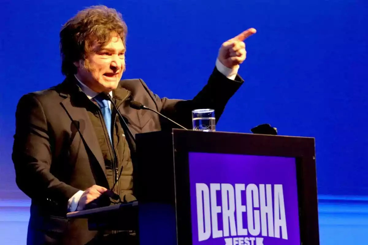 Hombre de cabello rizado y traje oscuro hablando en un podio con el cartel que dice DERECHA FEST y fondo azul